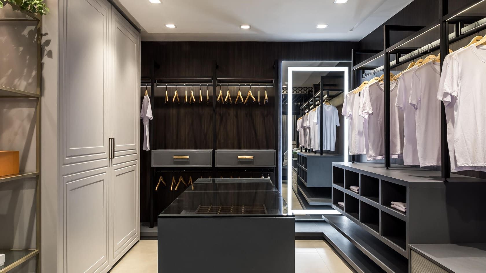 Walk-in closet with dark open wardrobes, a glass-top island and a lit mirror